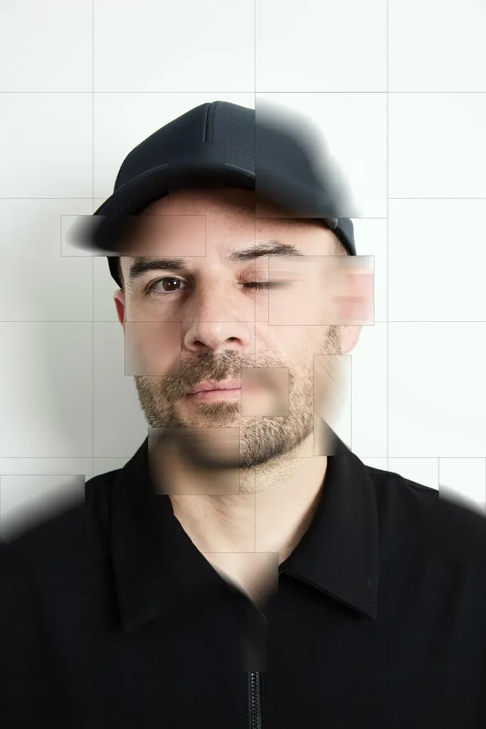 Colour portrait of a man in a black cap and black shirt facing the lens, his face recomposed in offset, blurred squares on a white grid ground.