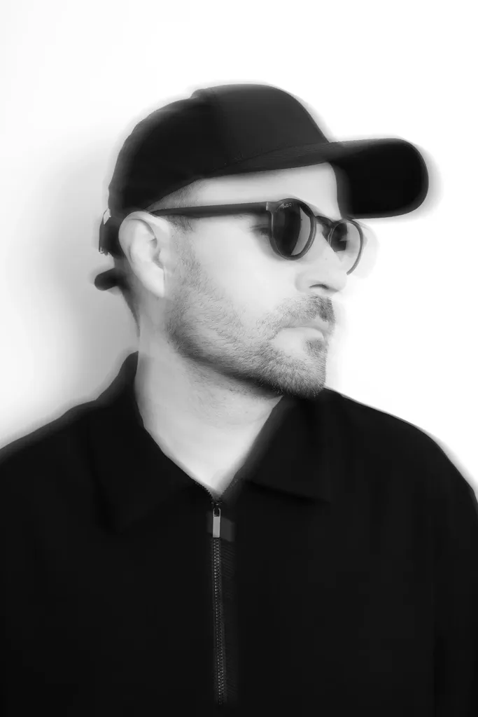 Black and white portrait of a man in a cap and sunglasses turned in profile to the right, his outline doubled by motion blur, on a white ground.