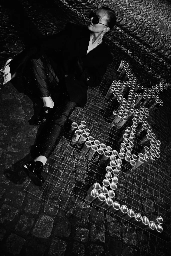 Black and white, seen from above: a person in a dark suit and sunglasses lying on cobblestones, next to letters formed by metal cylinders.