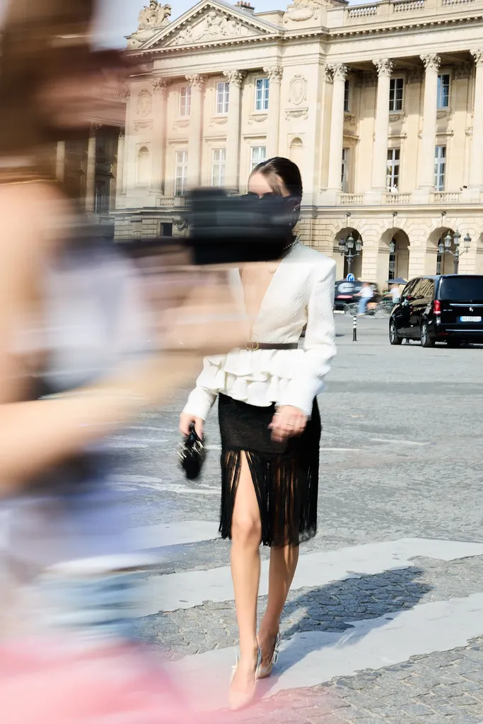 Colour, in the street: a woman in a white ruffled jacket and black fringed skirt crosses a pedestrian crossing, blurred figures in the foreground.