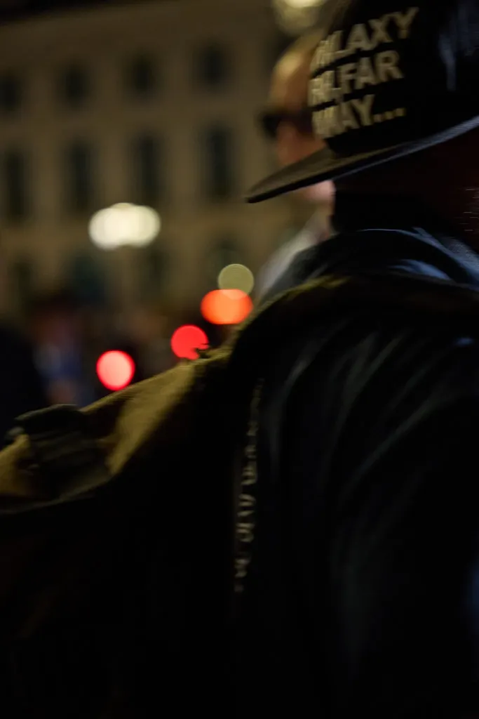 Colour, at night: from behind, a person in a dark jacket and embroidered cap, in front of the blurred lights of a street.