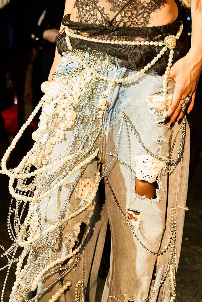 Colour, close-up at the waist: ripped jeans covered with strings of pearls and chains, under a black lace top.