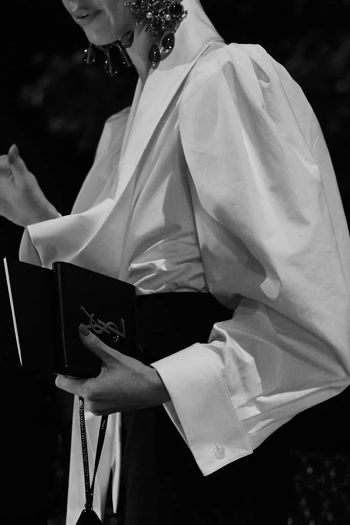 Black and white: the torso of a person in a loose white shirt with wide sleeves, holding a small black clutch, drop earrings.