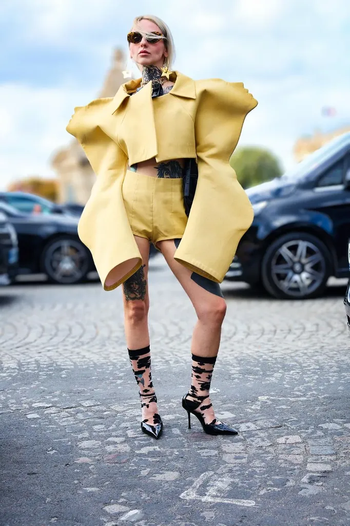 Colour, full length in the street: a woman in a yellow outfit with sculpted shoulders, black socks and pumps, in front of parked cars.