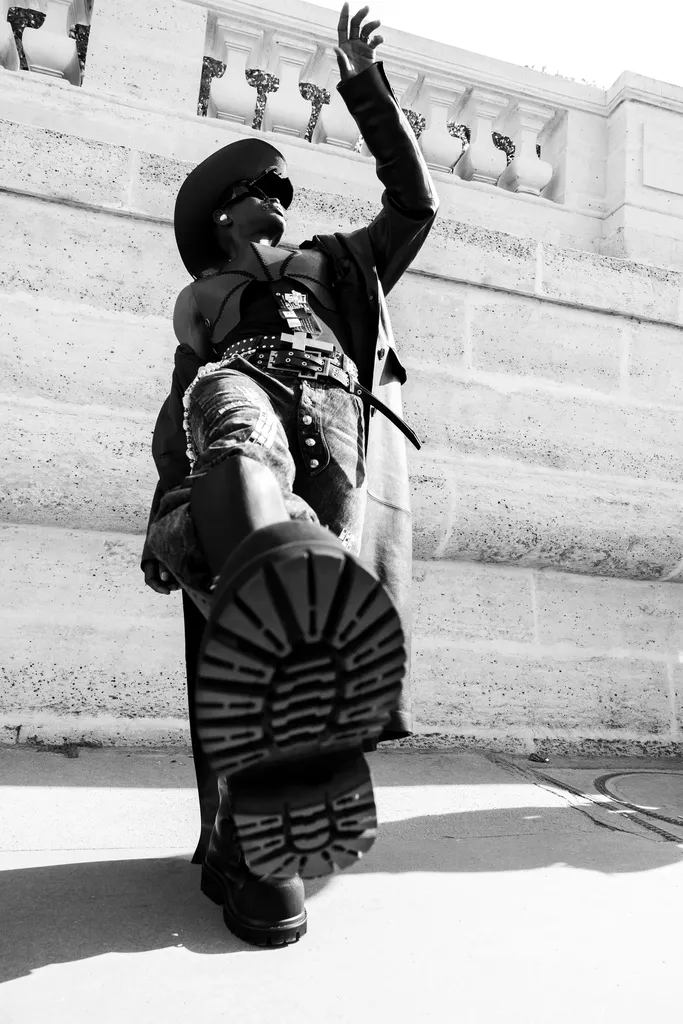 Black and white, low angle: a person in a hat and heavy boots raises a lug sole toward the lens, in front of a stone wall.