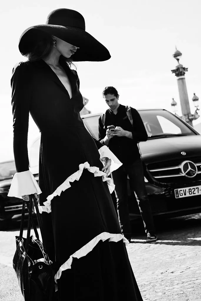 Black and white, in full sun: a woman in a long black dress with white ruffles and a wide-brimmed hat, in front of a black car.