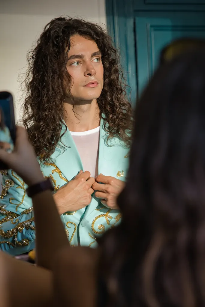 Colour: a young man with long curly hair, in a turquoise jacket embroidered with gold, surrounded by phones taking his picture.