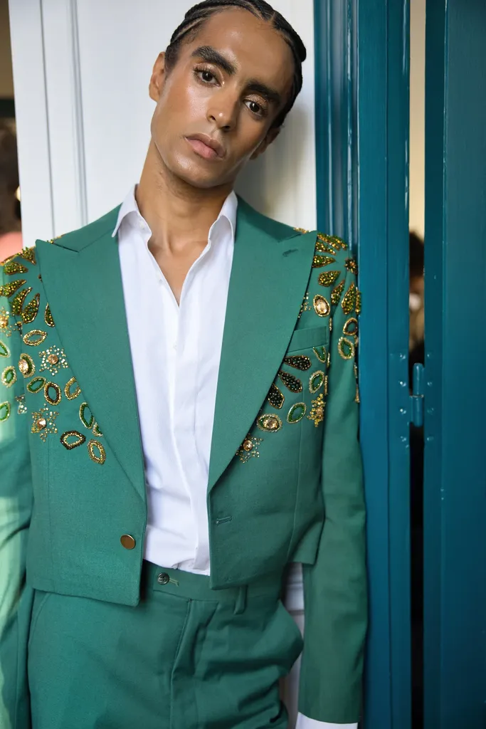 Colour, half-length portrait: a man in a green suit embroidered with sequins, open white shirt, leaning against a teal door.