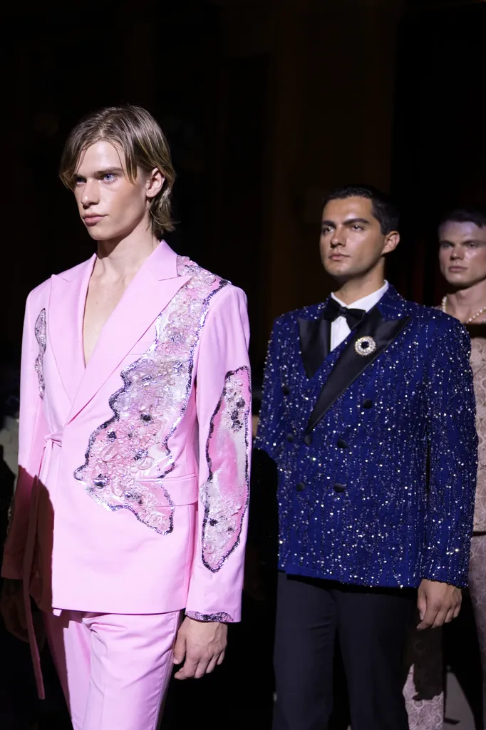 Colour, on a dark runway: two men in suits, one pink and embroidered, the other blue and sequined with a bow tie.