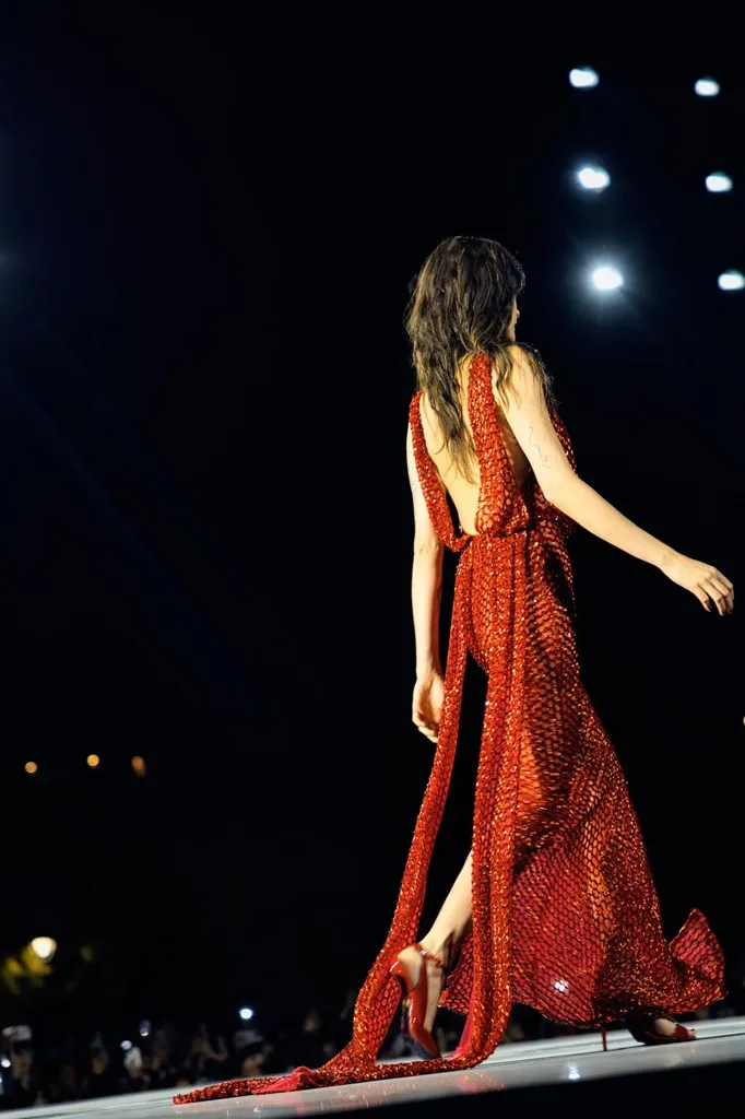 Colour, night runway: from behind, a model in a long, backless, sparkling red gown walks toward the spotlights.