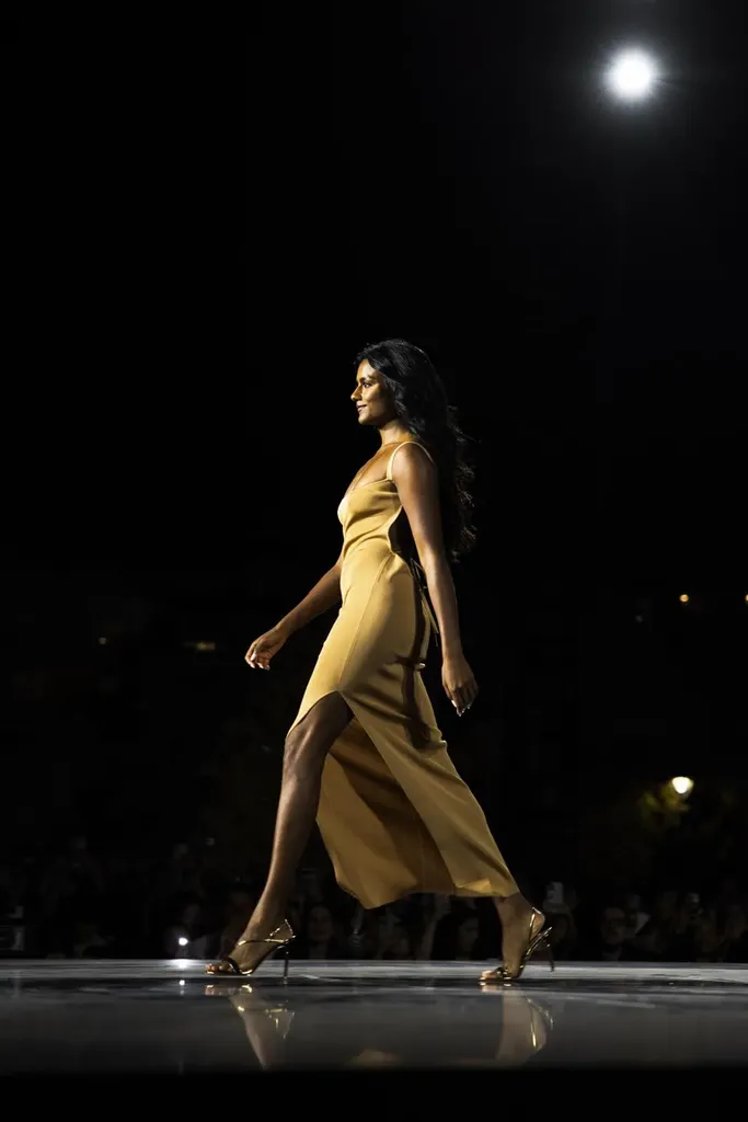 Colour, night runway: a model in a golden satin dress walks on a glossy runway, a spotlight above her.