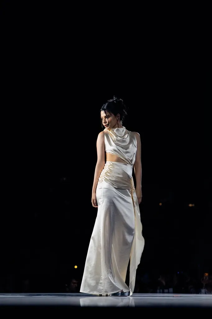 Colour, night runway: a model in an ivory satin top and long skirt, eyes lowered, alone in the light.