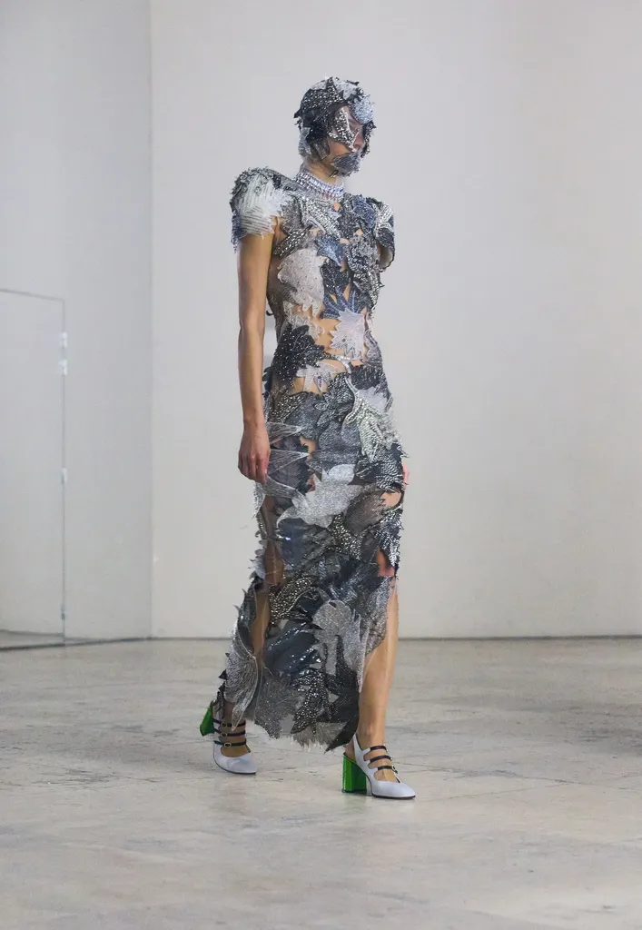 Colour, presentation in a bright room: a model in a long dress covered with silver and grey pieces, her face masked.