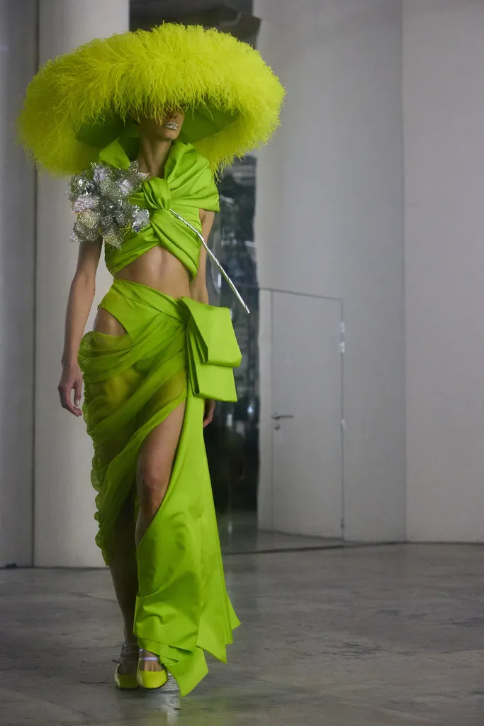 Colour, presentation in a bright room: a model in a draped lime green outfit, wearing a huge feathered hat.