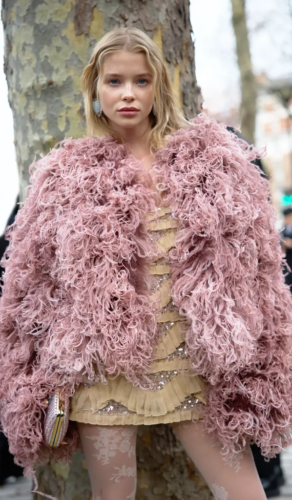 Colour, in the street: a young blonde woman in a dusty pink feather coat and golden dress, in front of a tree trunk.