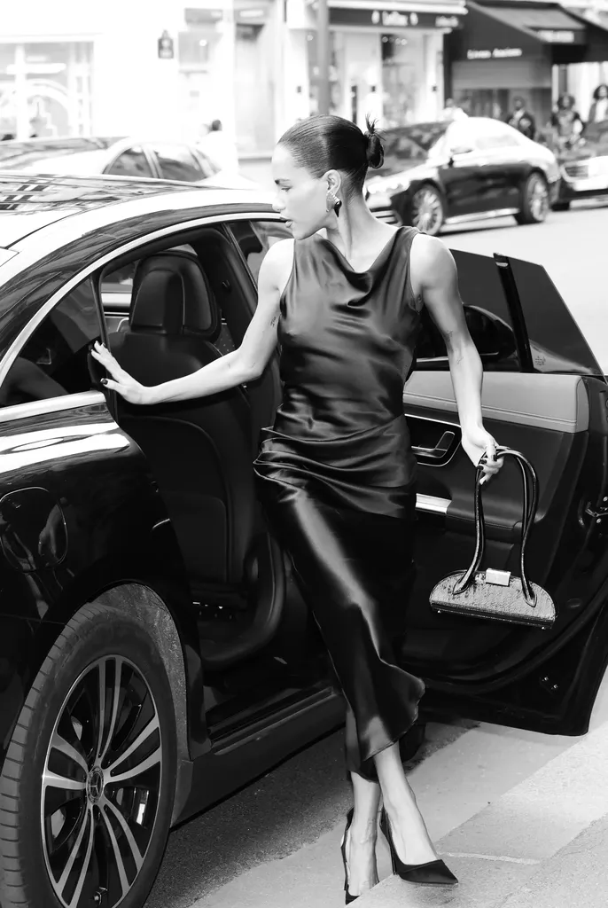 Black and white: a woman in a dark satin dress steps out of a car, door open, bag in hand.