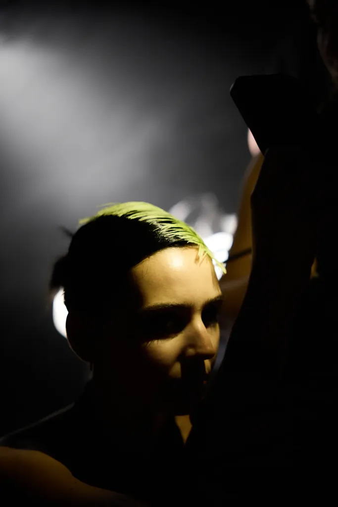Colour, backstage: the face of a person with acid green dyed hair, caught in low raking light on a black ground.