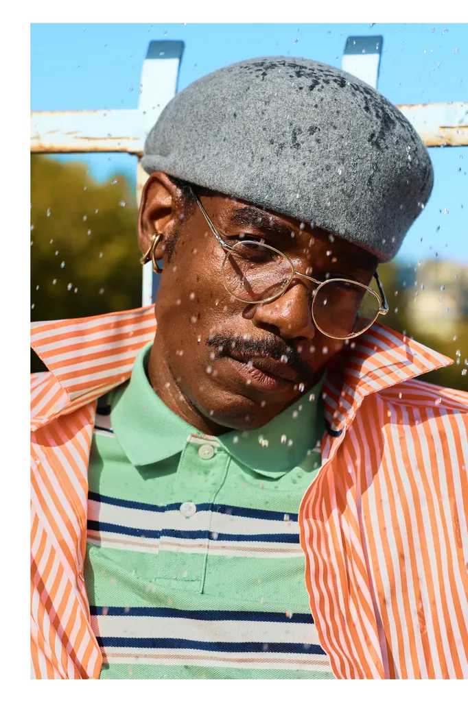 Colour, outdoor close-up: a man in a grey beret and round glasses, orange striped shirt over a green polo, under drops of water.