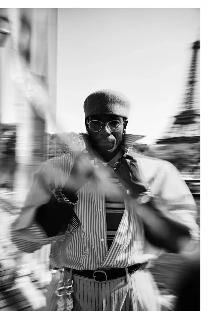 Black and white, motion blur: a man in a beret and round glasses, striped shirt, adjusts his collar in the street.