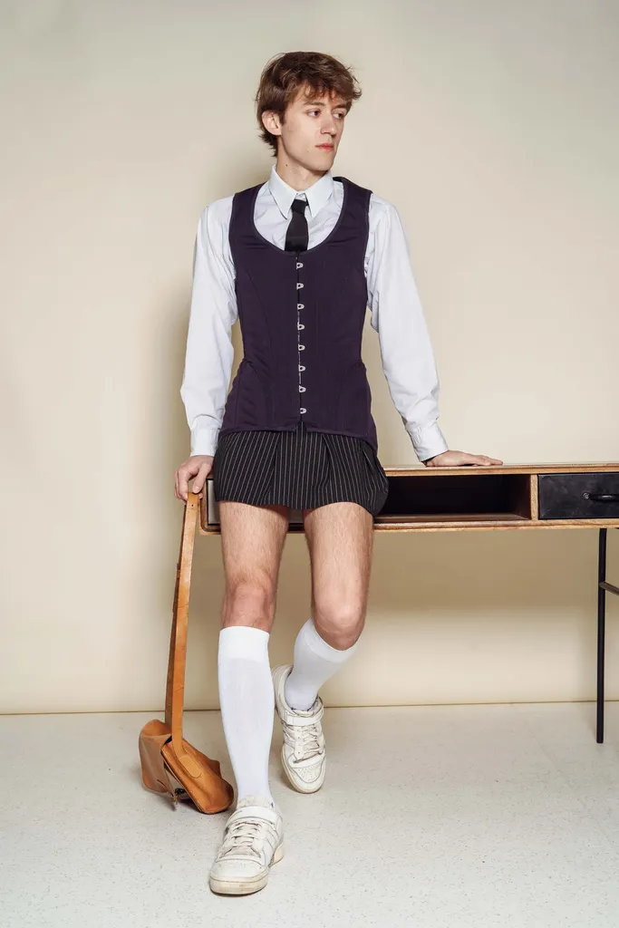 Colour, studio on a beige ground: a young man in a white shirt, dark waistcoat, shorts and knee socks, leaning against a desk.
