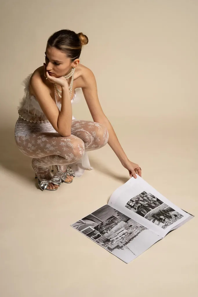 Colour, studio on a beige ground: a young woman crouching in pale lace turns the pages of a magazine on the floor.