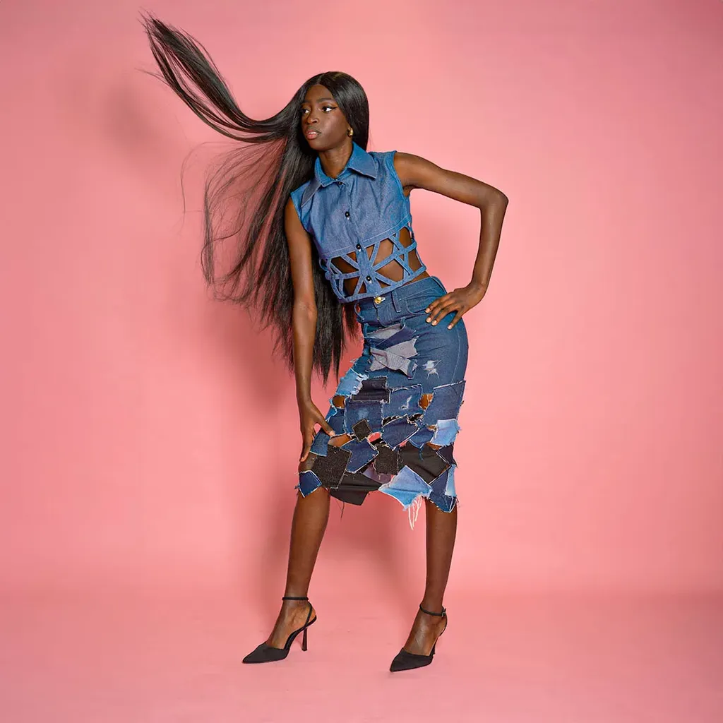 Colour, studio on a pink ground: a woman in a patchwork denim outfit, long hair flying, hand on her hip.