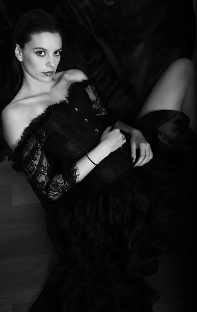 Black and white: a woman lying down in an off-the-shoulder black lace dress, looking at the lens.
