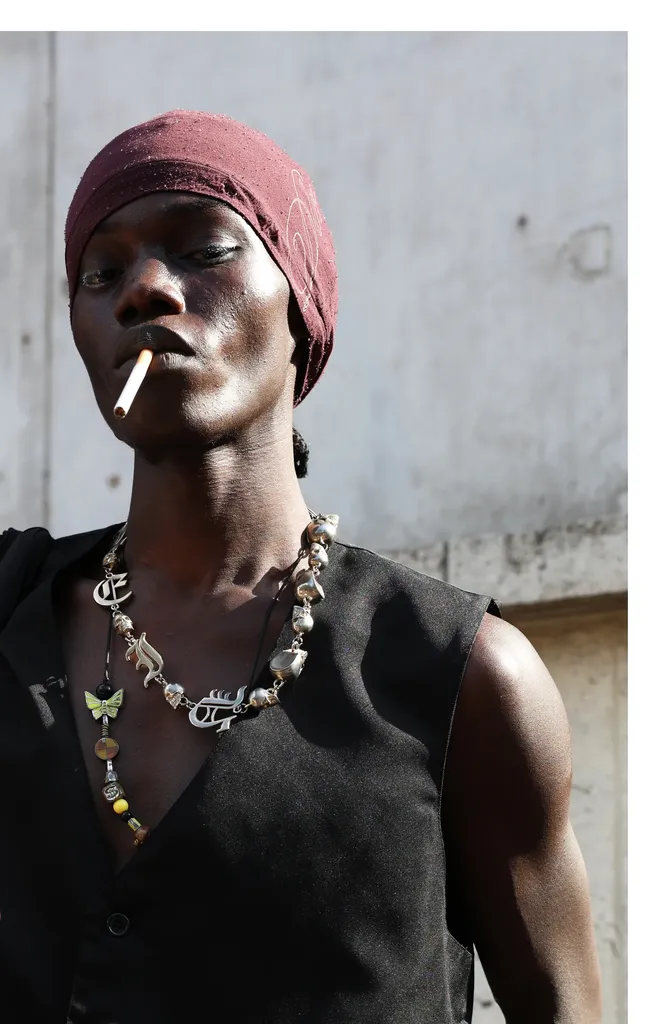 Colour, daylight portrait: a man in a burgundy headscarf and black waistcoat, cigarette in his mouth, necklaces of beads and charms.