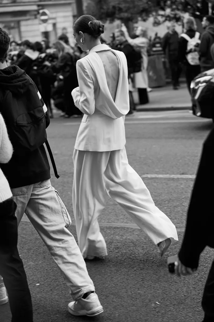 Black and white, in the street: from behind, a woman in a pale trouser suit with an open back crosses among passers-by.