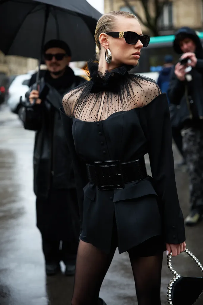 Colour, in the rain: a woman in black sunglasses, a dotted tulle collar and a belted jacket, surrounded by photographers.