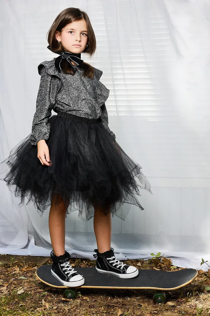 Full-length portrait of a girl standing on a skateboard in a glittery top, black tulle skirt and sneakers, in front of a white curtain, in colour.