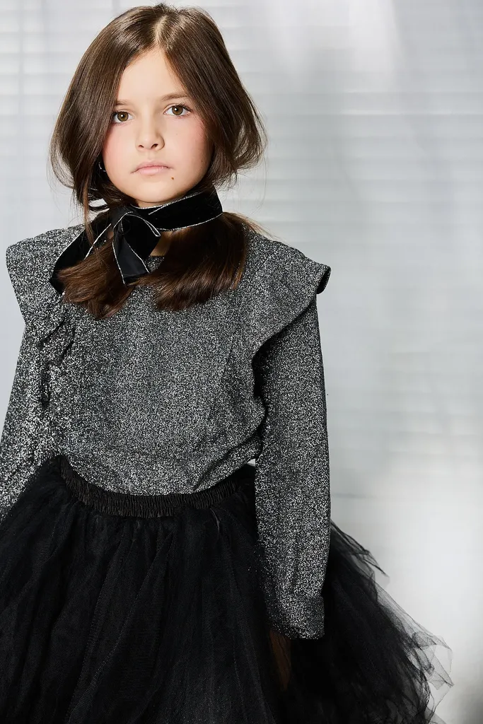 Portrait of a girl in a glittery top with a black bow at the collar, looking straight at the camera, in front of a white curtain.