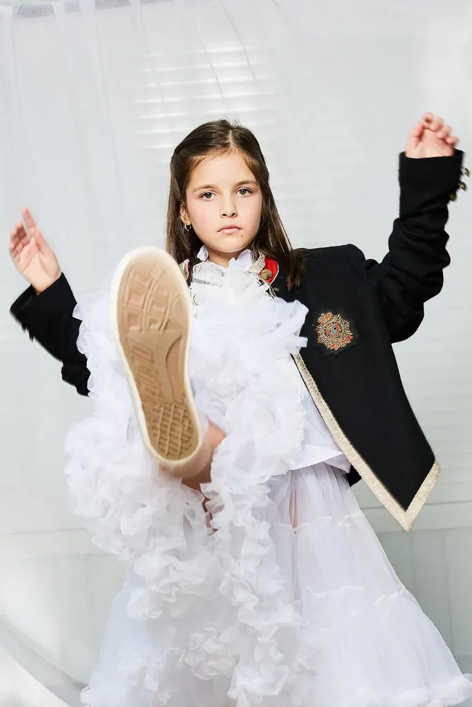 Girl in a white petticoat and embroidered black jacket, one leg raised toward the camera, the sole of her sneaker in the foreground.