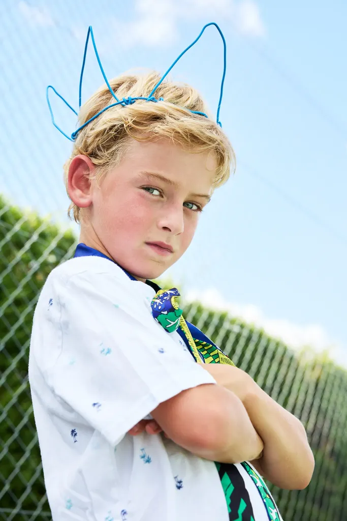 Blond boy with crossed arms, a blue wire headband, white shirt and printed tie, in front of a wire fence and blue sky.