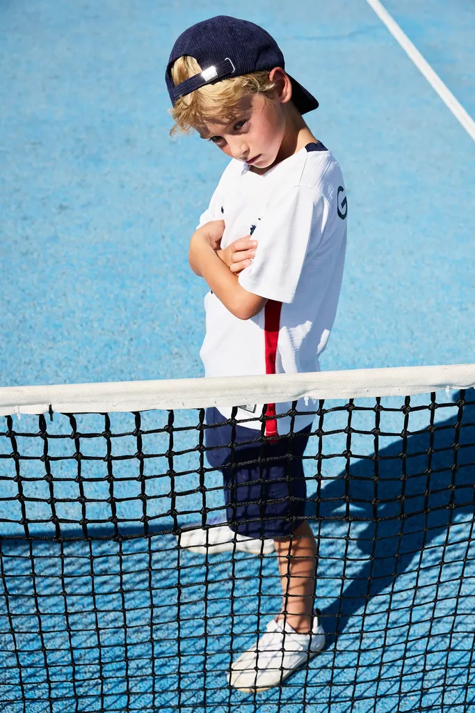 Boy in a cap and white T-shirt by the net of a blue tennis court, head lowered, in colour.