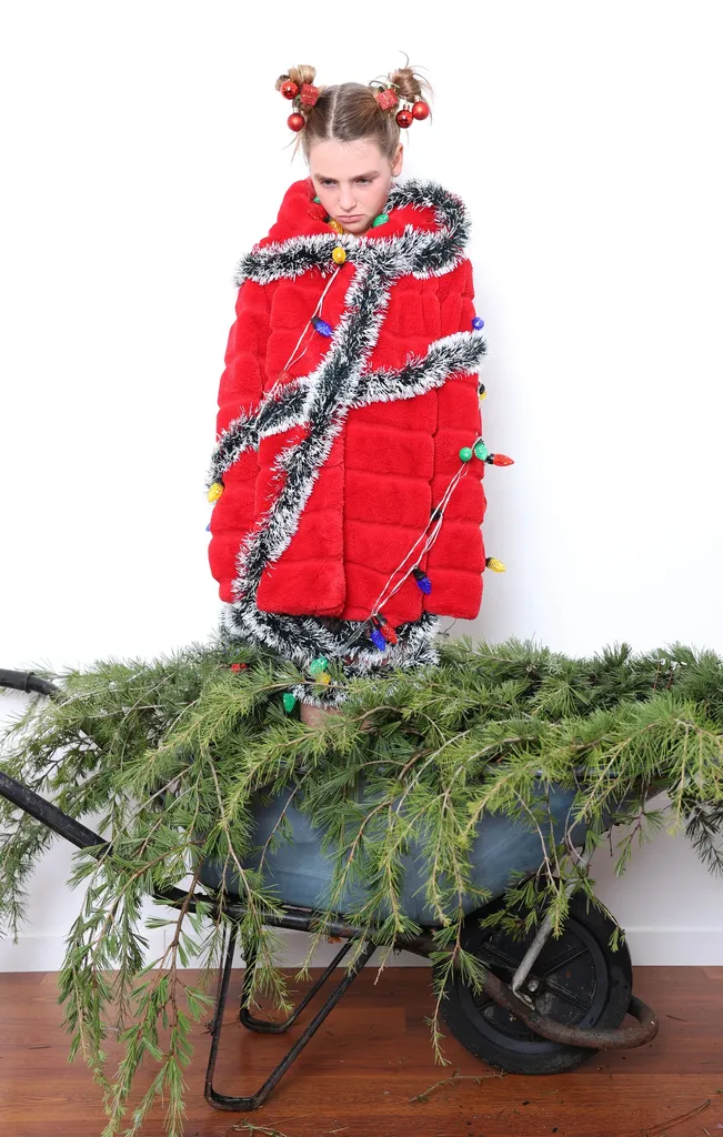Girl standing in a wheelbarrow full of fir branches, red coat wrapped in silver tinsel, on a wooden floor.