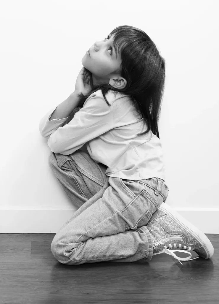 Girl kneeling against a white wall, face turned upward, on a wooden floor, in black and white.
