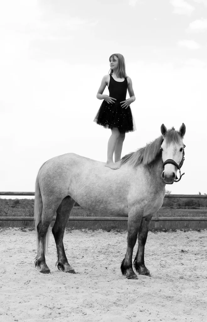 Girl in a black tutu standing on the back of a pony in a sand arena, in black and white.