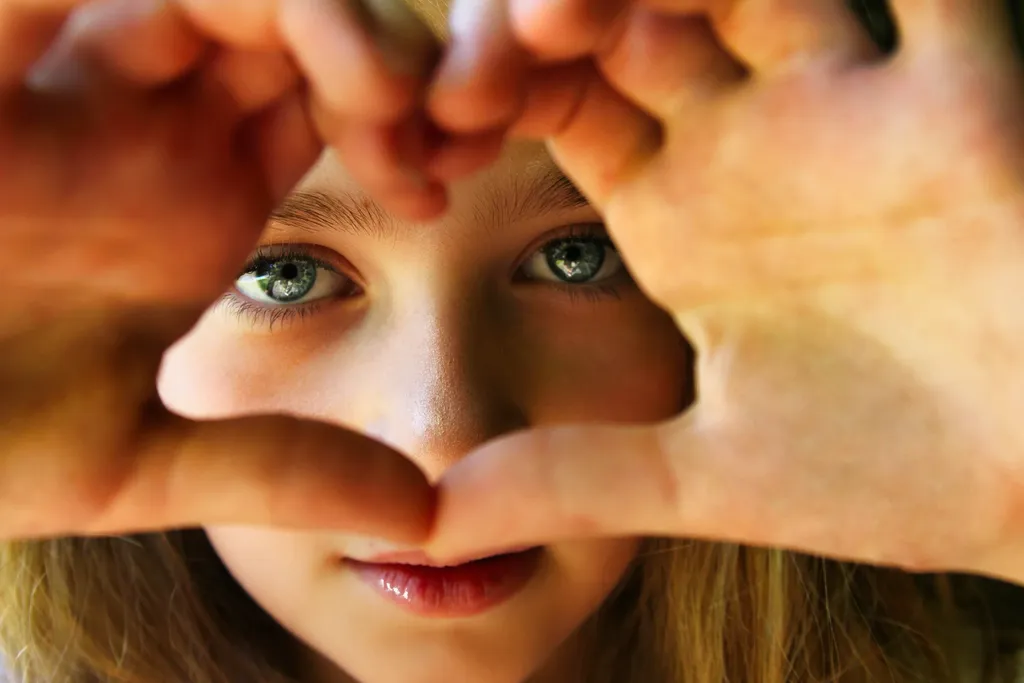 Close-up of green eyes framed by hands making a heart shape, in colour, warm light.