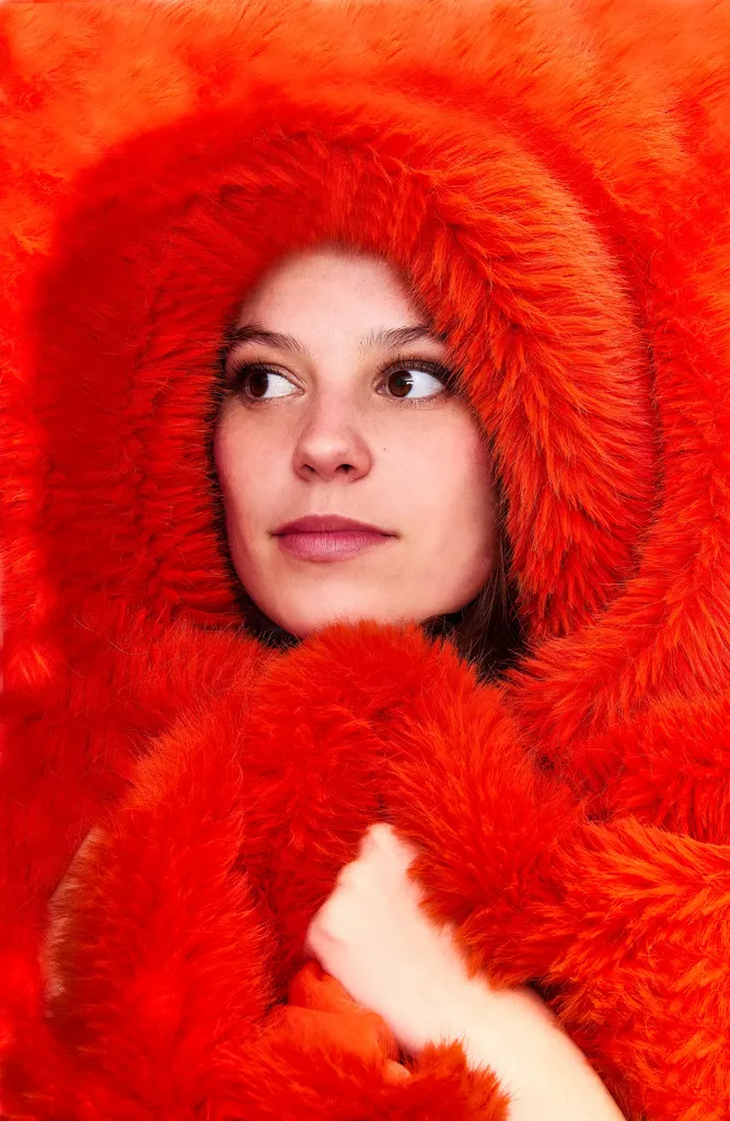 Colour portrait of a young woman wrapped in a bright red fur hood, looking to the side, one hand near her chin.