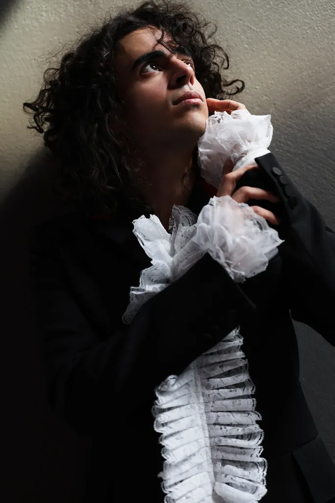 Dark-toned portrait of a young man with long curly hair and a white ruffled jabot, hand on his chest, looking up.