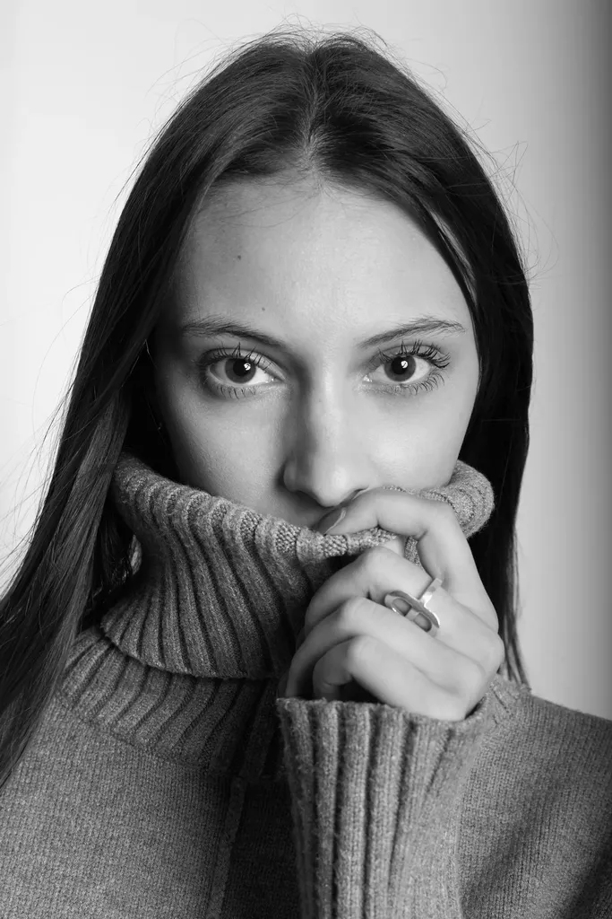 Black and white portrait of a young woman with long hair, her polo neck pulled up over her mouth, hands joined.