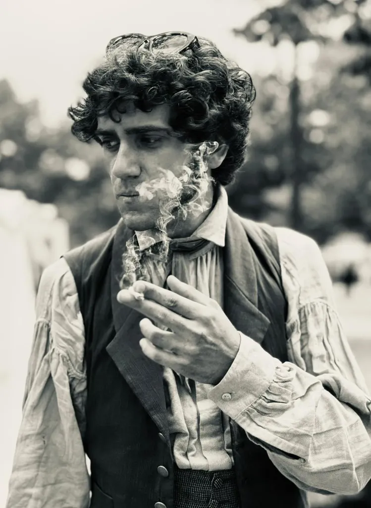 Black and white photograph of a young man with curly hair in period costume, waistcoat and loose shirt, smoke in front of his face.