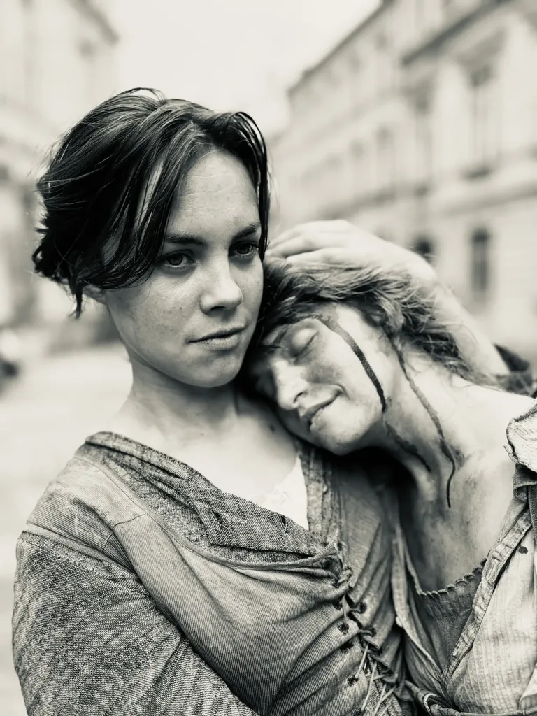 Black and white photograph of two women in period costume in a street, one resting her head on the other's shoulder.