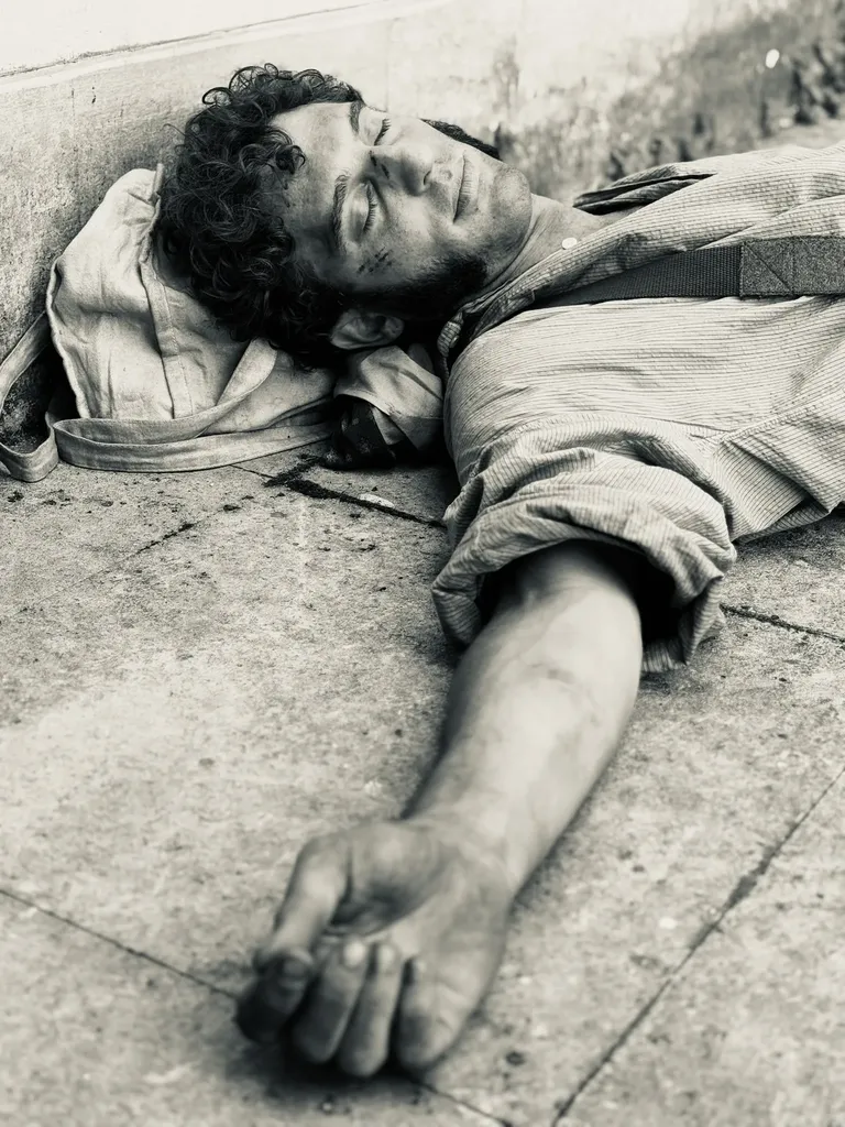 Black and white photograph of a man in period costume lying on paving stones, eyes closed, one arm stretched out.