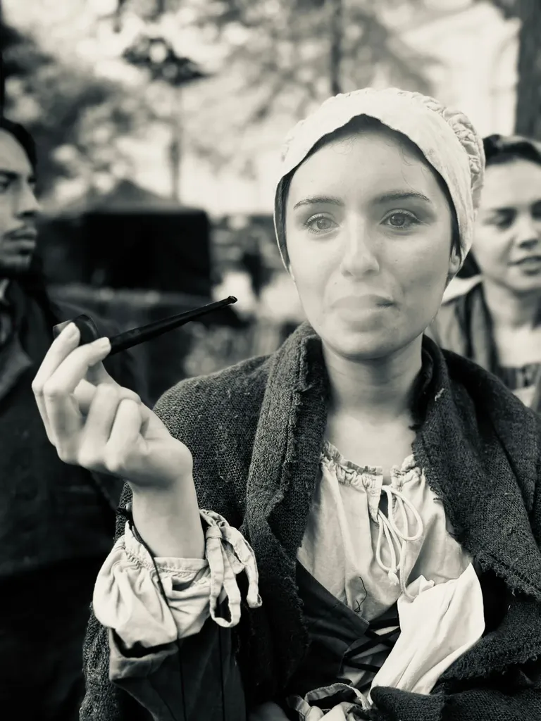 Black and white photograph of a young woman in a bonnet and period costume holding a long pipe, other people in costume around her.