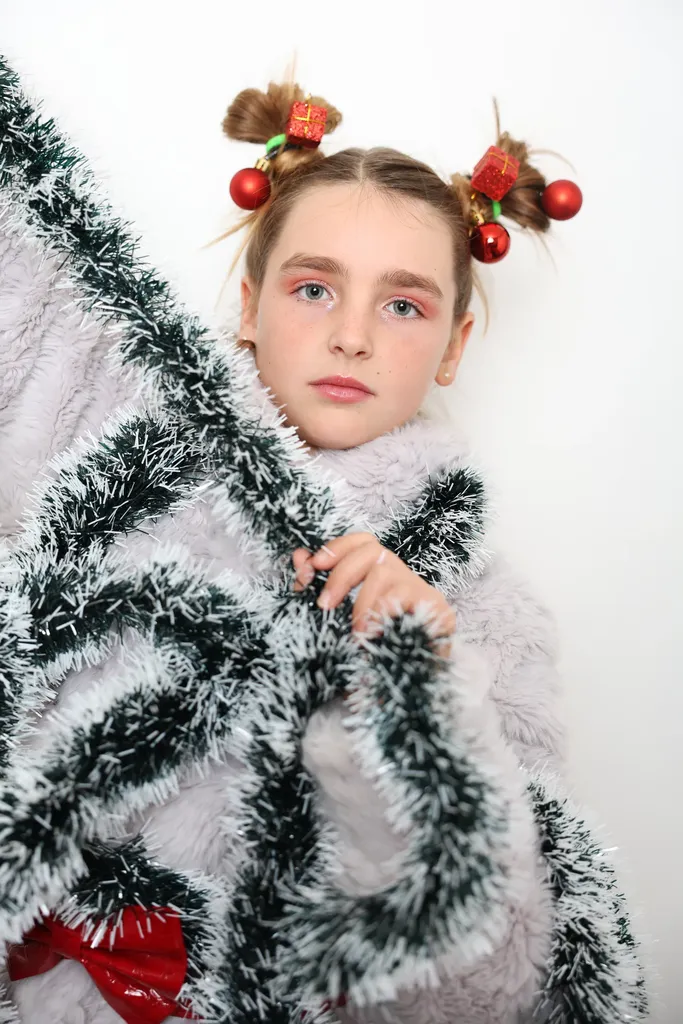 Colour portrait of a girl with two hair buns trimmed with red baubles, wrapped in tinsel and white fur, on a white background.