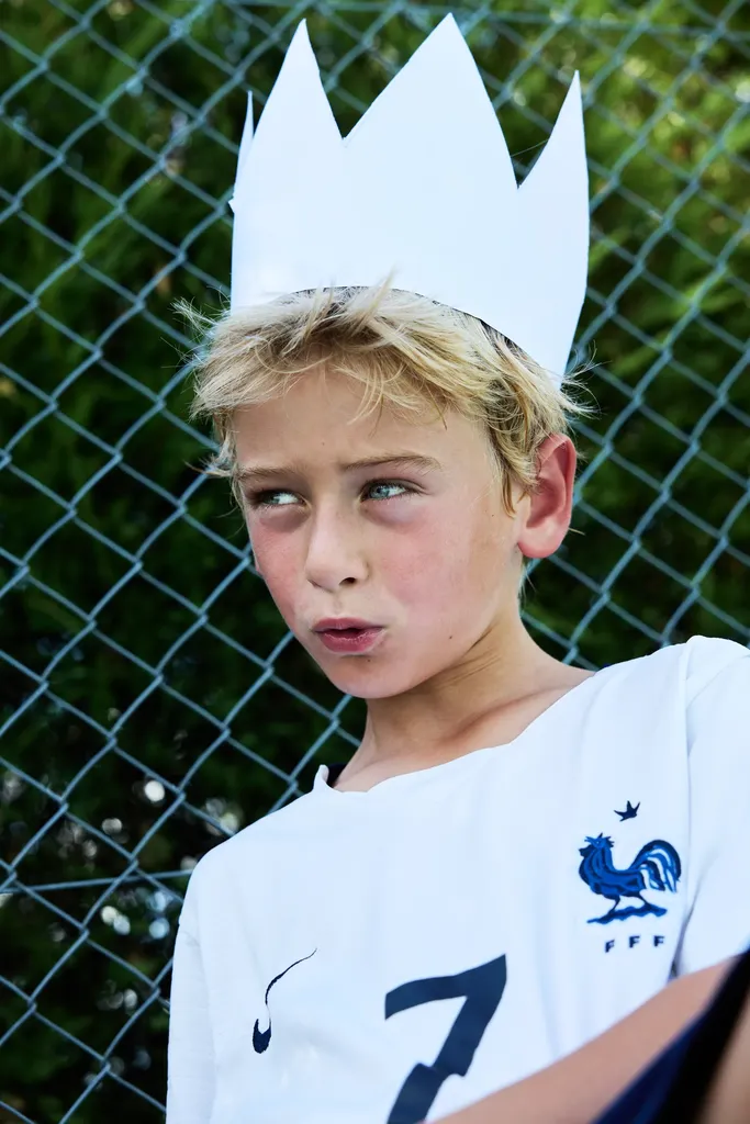 Colour portrait of a fair-haired boy wearing a white paper crown and a white football shirt, in front of a wire fence.